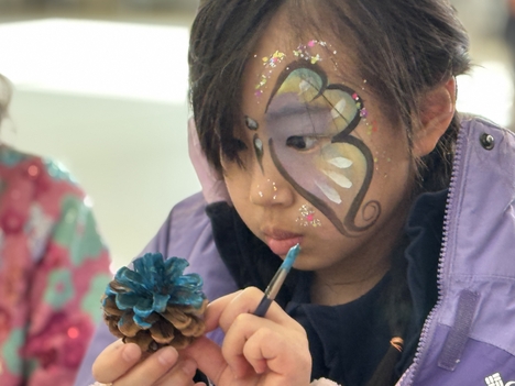 A young girl with butterfly facepaint studies a pinecone she is painting blue.