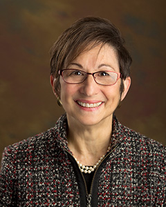 A headshot of a woman with brown hair wearing a multi-color jacket smiling at the camera.