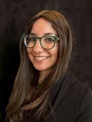 A head-and-shoulders shot of a woman with long brown hair and glasses smiling at the camera. 