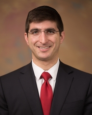 A head-and-shoulders shot of a man with short black hair wearing a black suit, white shirt and red tie smiling at the camera.