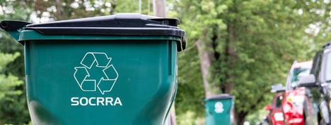 A green recycling container with a black lid, waits at the curb.