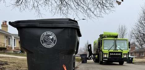 A waste bin stands in a street while a garbage truck approaches.
