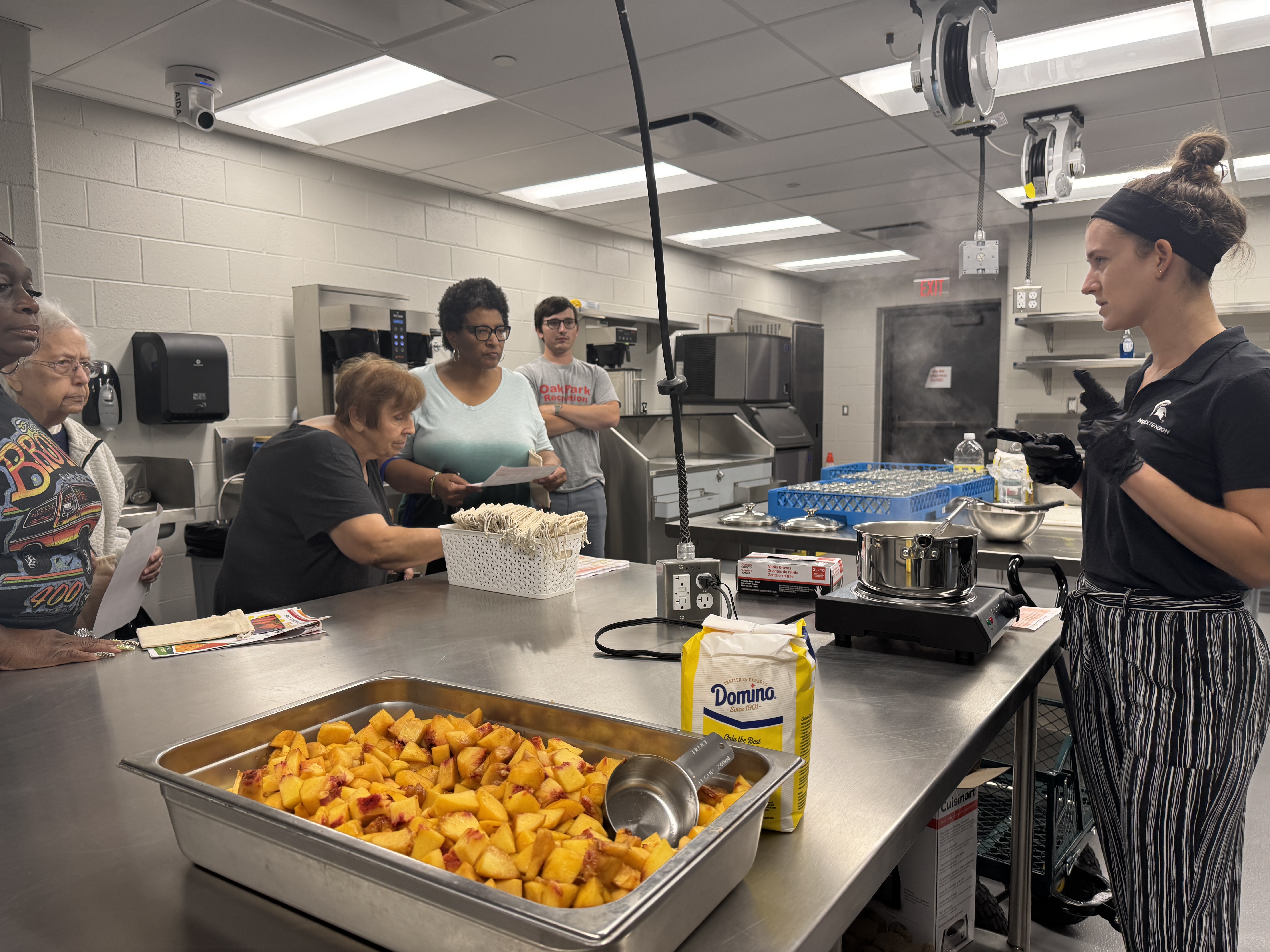 A group watches a cooking demonstration in a commercial kitchen.