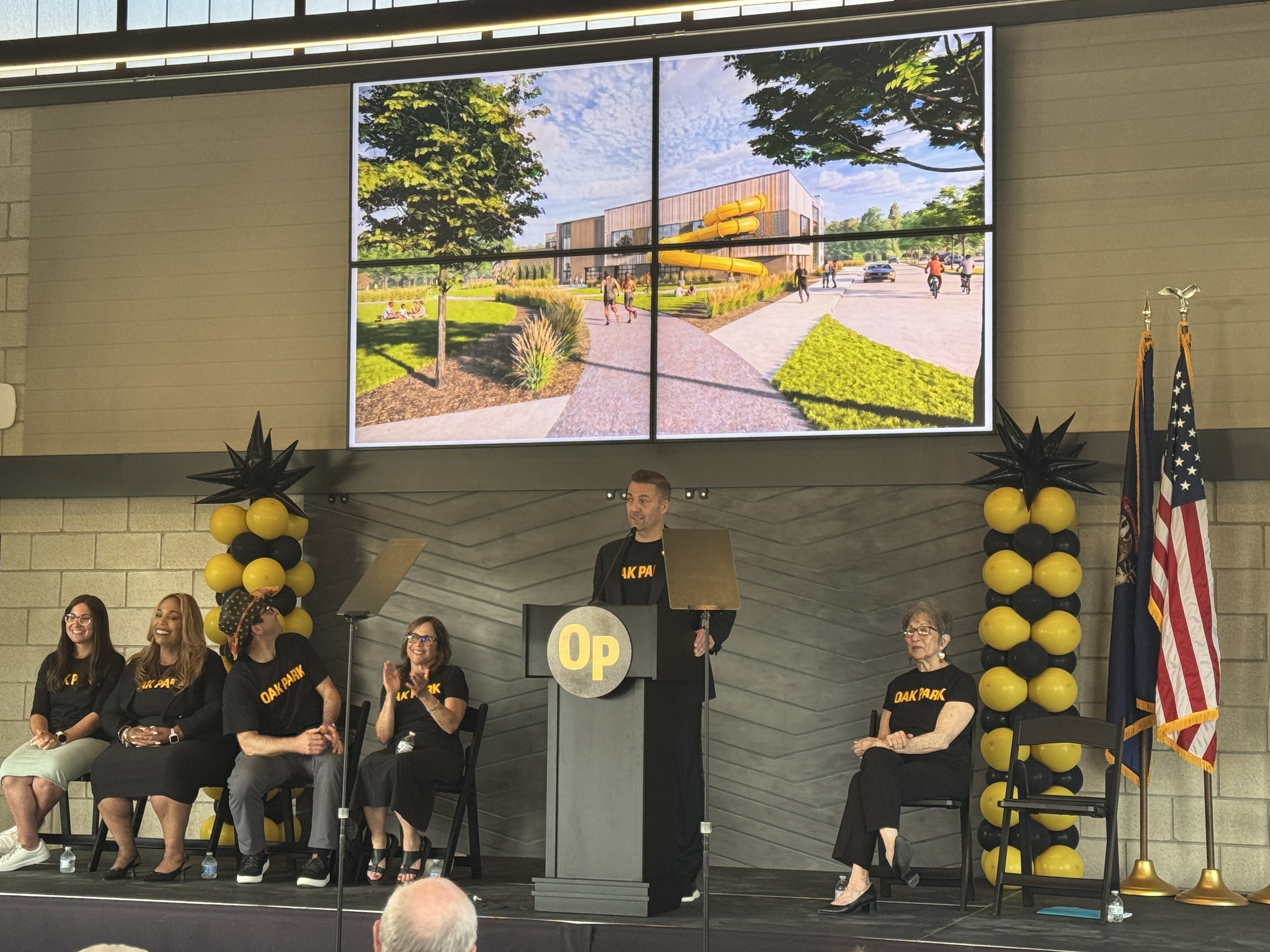 Six people on a stage with a large screen television and gold and black balloons in the background.