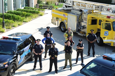 A group of Oak Park Michigan Department of Public Safety officers stand in a circle formed by two police vehicles and a fire truck.