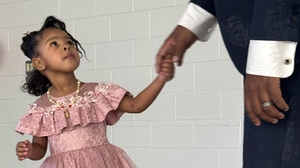 A young girl in a pink party dress holds hands and look up at a large man.
