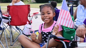A young girl in a colorful outfit holds an american flag while sitting in a lawn chair smiling at the camera.