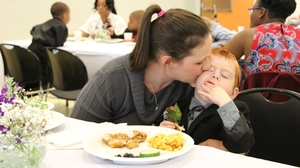A woman kisses a redheaded child sitting at a banquet table with a plate of food in front of them.