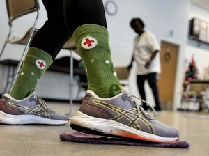 A pair of feet wearing athletic shoes and green socks with a red cross inside a white circle.