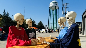 Four Halloween skeltons wearing clothing are seated at a table playing cards with a water tower in the background.