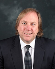 A head and shoulders shot of a man with brown hair in a black suit and white shirt.