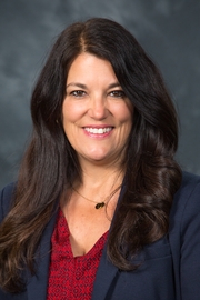 A headshot of a woman with long black hair wearing a red blouse and black jacket smiling at the camera.