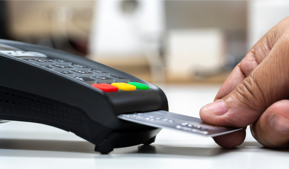 A hand holds a credit card near a payment processing terminal.