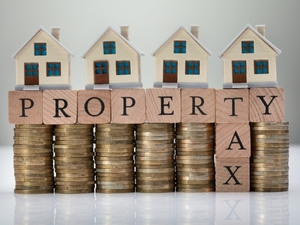 Four toy houses sit on top of wood tiles letters that spell out property tax anad are being help up back stacks of coins.